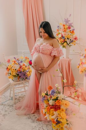 Maternity photoshoot with pink dress and colorful floral arrangements in bright studio setting.