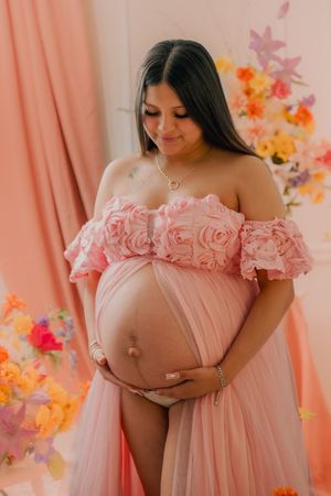 Maternity photoshoot featuring a pink off-shoulder dress with floral decorations and peach curtains in the background.