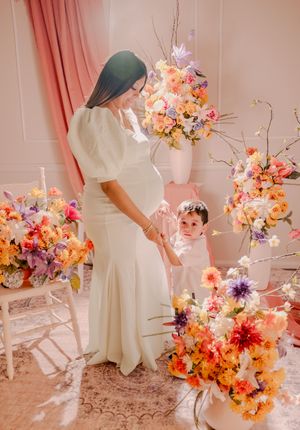 White dress maternity session with bright floral arrangements against pink curtains.