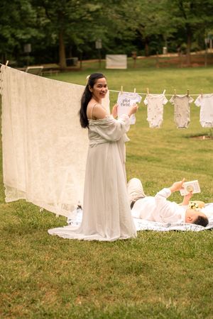 Woman in flowing white dress smiles while hanging laundry on a line outdoors.