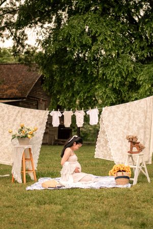 Rustic maternity photo setup with white linens, baby clothes, and flowers in an outdoor garden setting.
