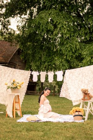 Garden maternity photo featuring white linens, hanging baby clothes, and rustic decorative elements.