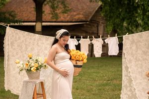 Expectant mother in white dress poses with sunflowers beside vintage laundry display.