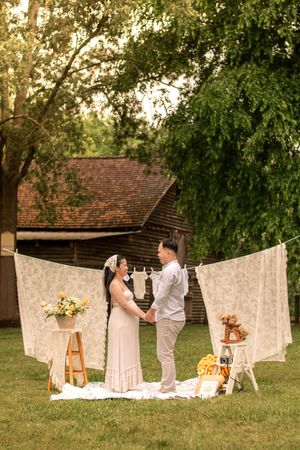 Maternity photography session at golden hour with white lace curtains and rustic props in a garden setting.