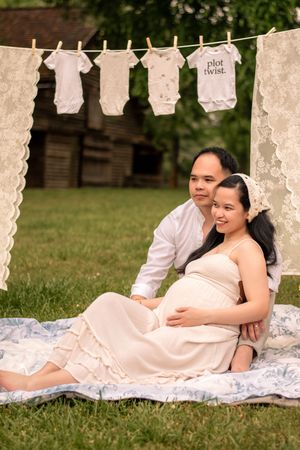 Expectant couple relaxing on white blankets during outdoor maternity session with hanging lace decorations.