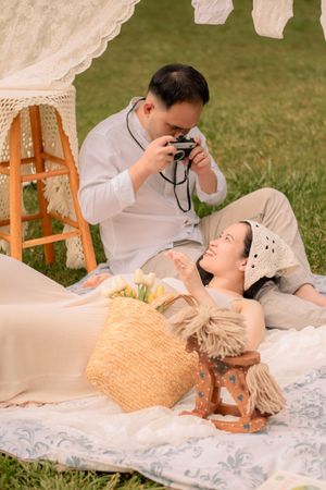 Couple lounging on white blankets during an outdoor maternity photoshoot with vintage camera props.