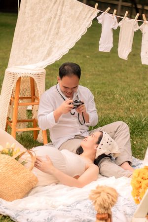 Intimate maternity photography session with vintage camera props and white lace backdrop outdoors.