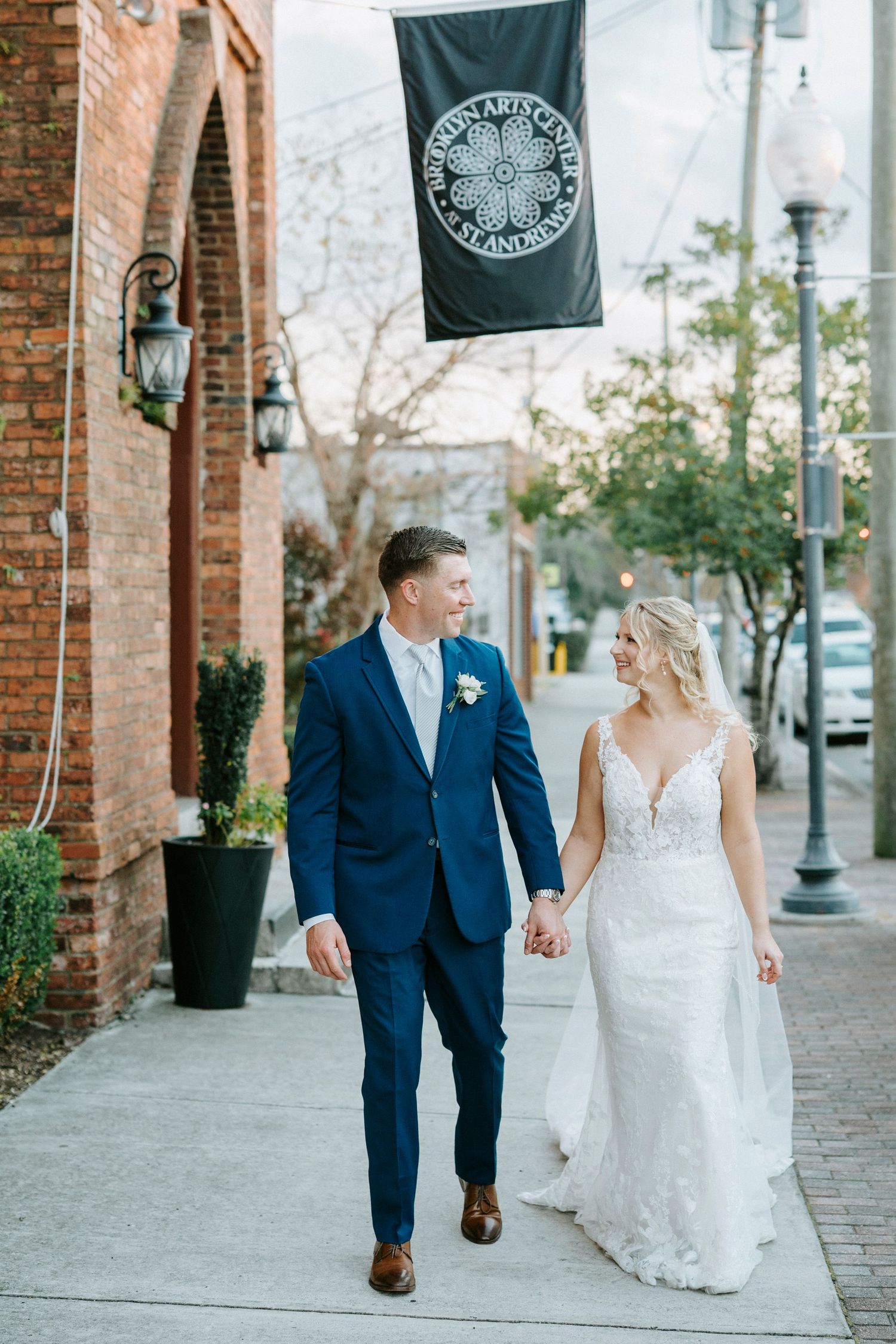 A couple in wedding attire walks together on a brick sidewalk under a green flag on a sunny day.