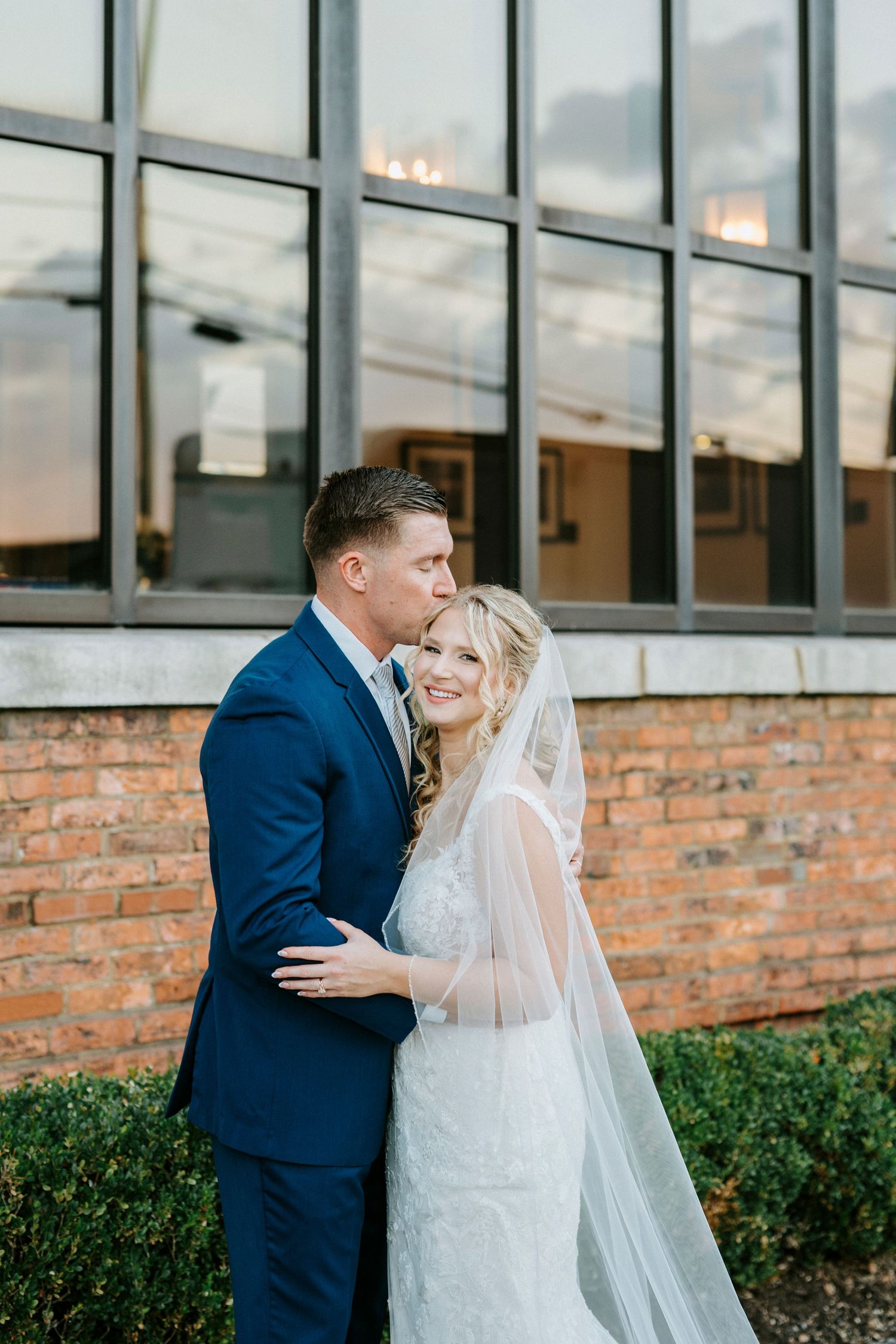 Bride and groom share an intimate moment against a brick wall with large windows at their wedding venue.
