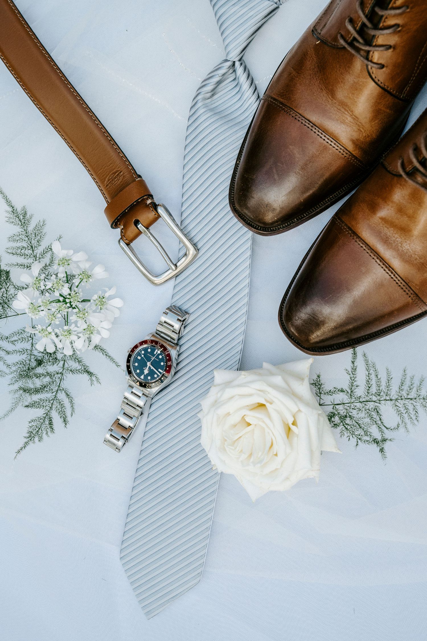 Wedding accessories including brown leather shoes, gray striped tie, silver watch, leather belt and white rose on light background.