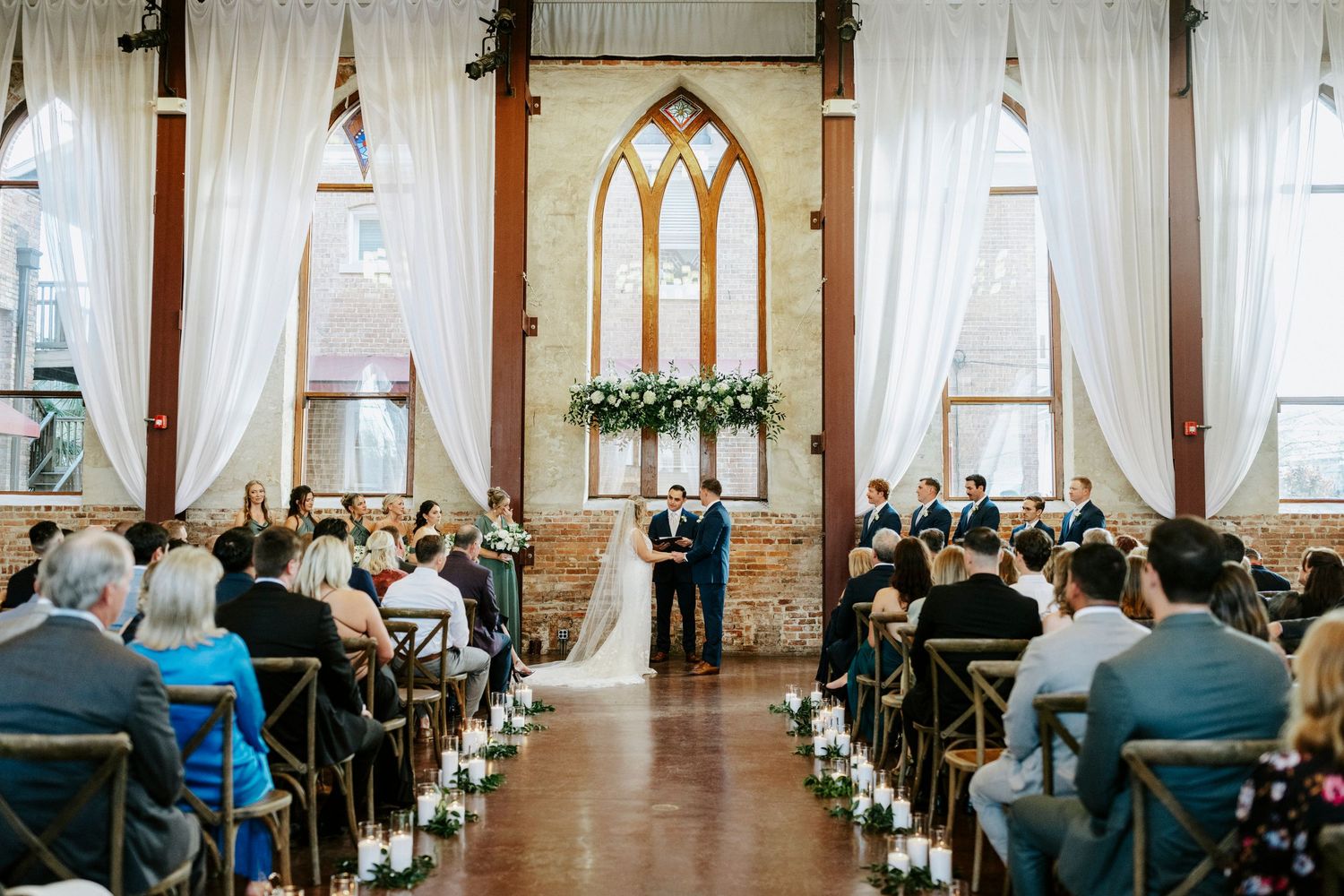 Wedding ceremony venue with elegant white drapes, arched windows, and rows of wooden chairs set up for guests.