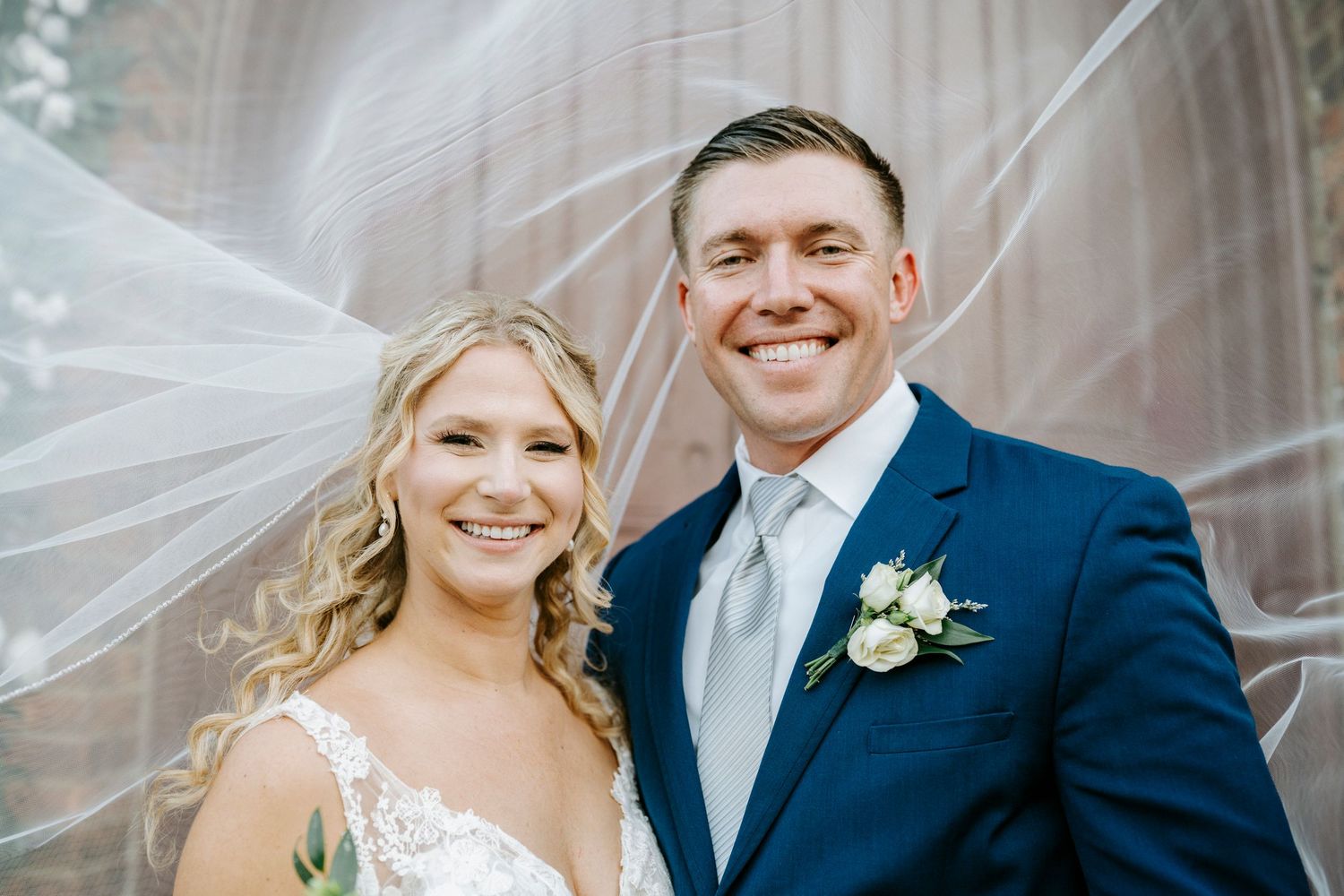 A happy couple poses together on their wedding day, with a flowing veil and navy suit with boutonniere.
