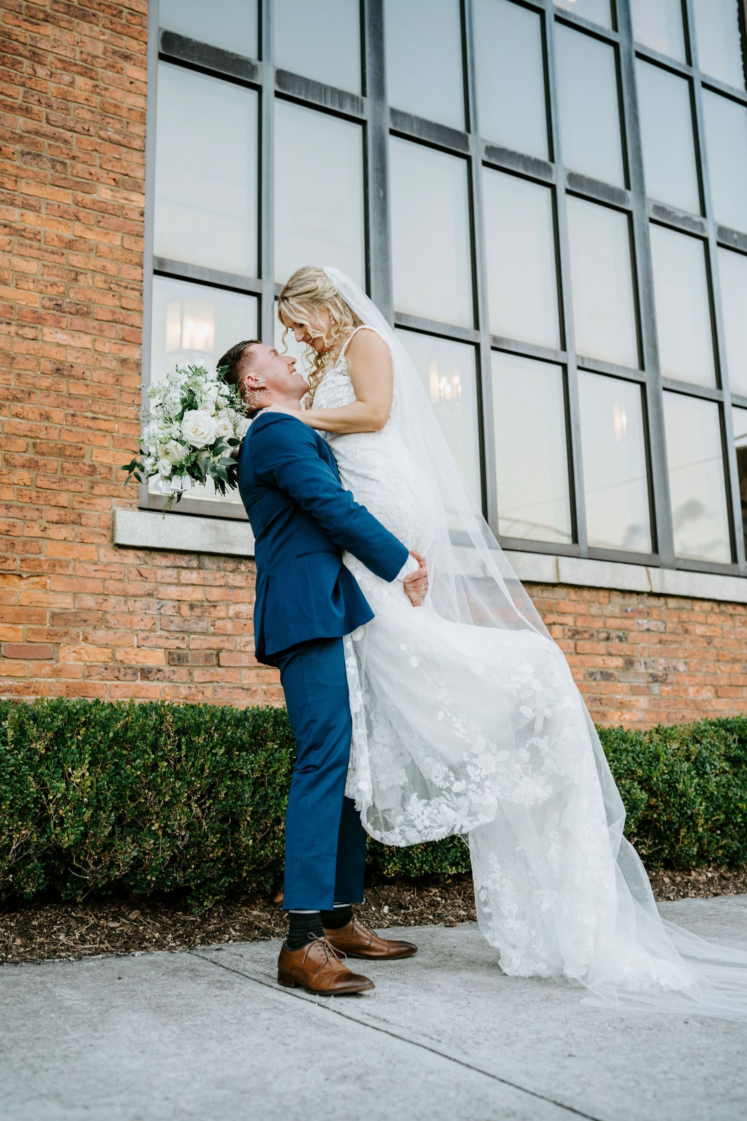 A romantic wedding moment outside a modern building with large windows and brick walls.