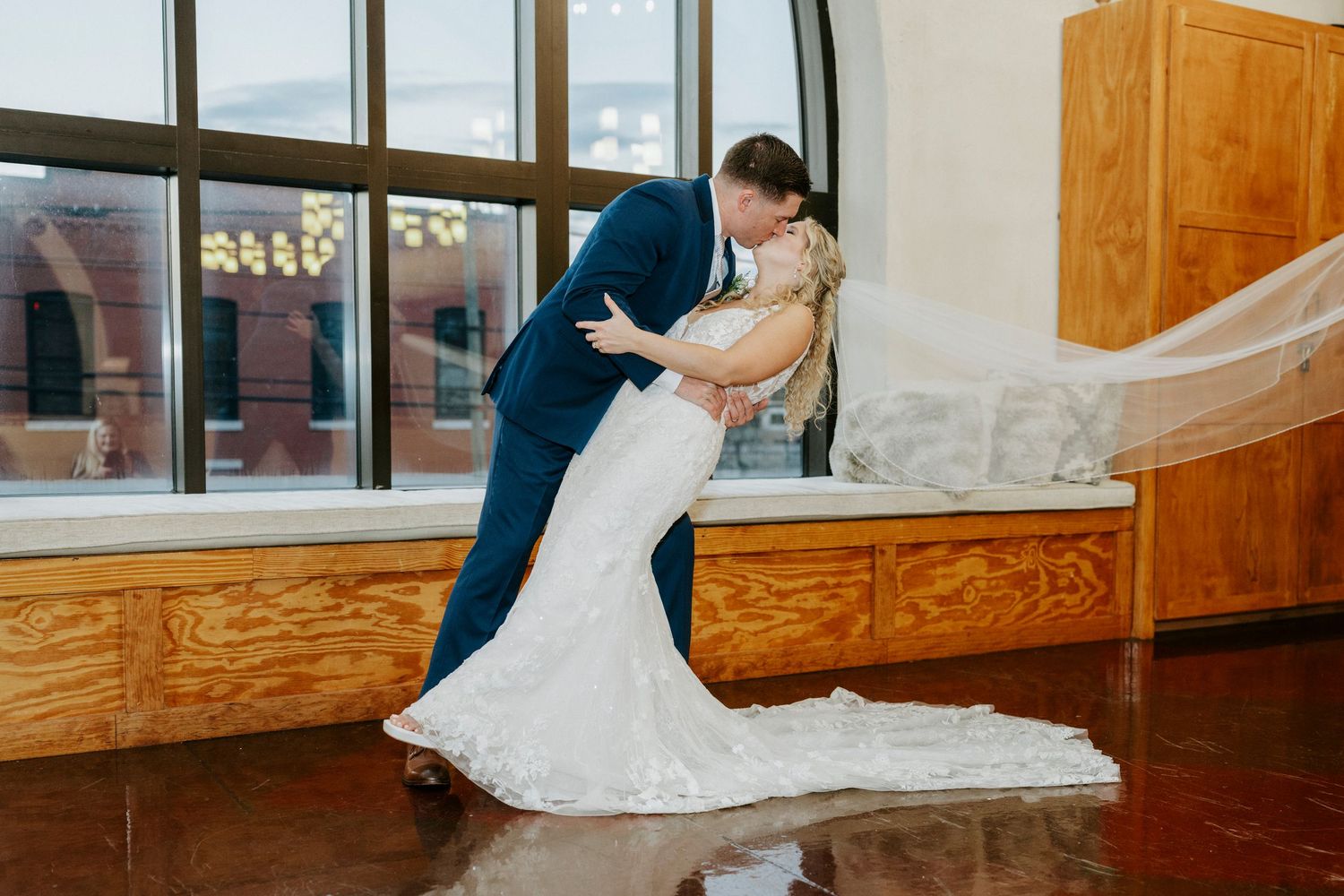 A romantic couple in formal wedding attire share an intimate dance dip pose in a sunlit room with wooden details.