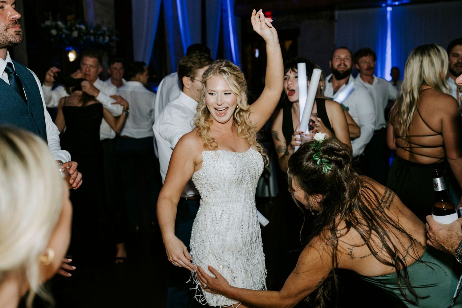 Wedding reception guests dancing and celebrating on a crowded dance floor with blue lighting in the background.