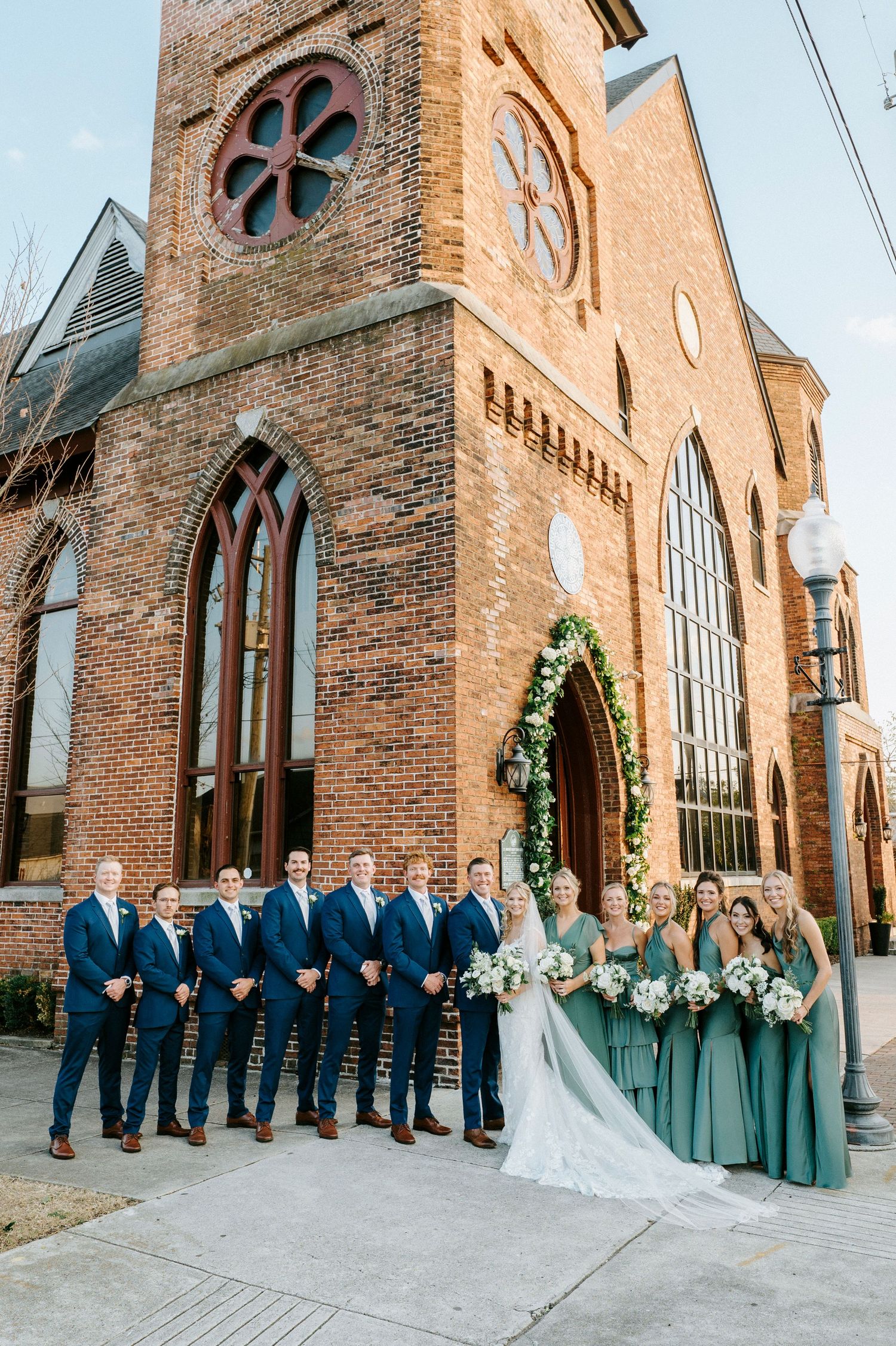 Wedding party in navy suits and sage green dresses pose together outside a historic brick church with arched windows.