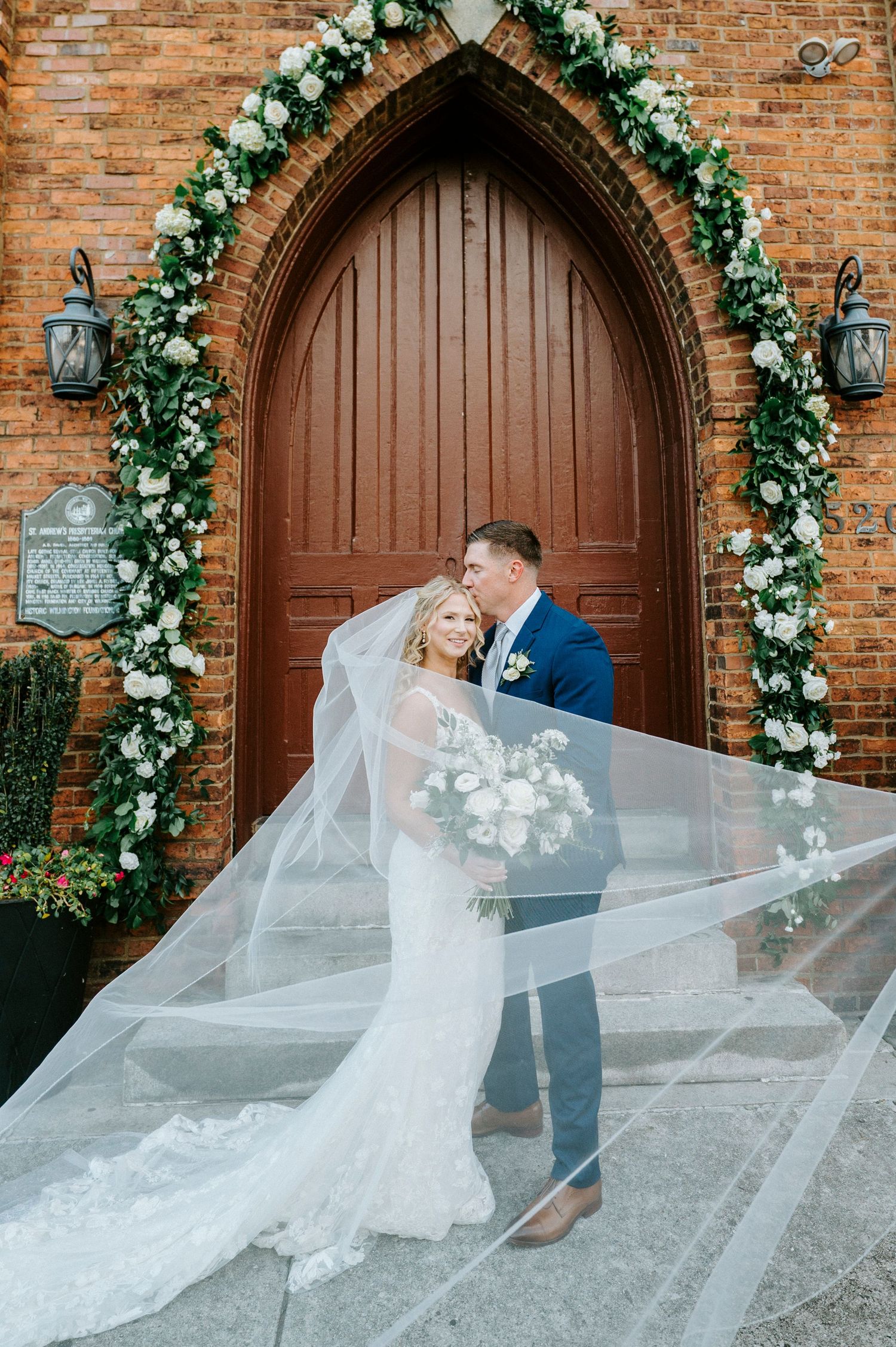 A couple shares a romantic moment under a floral arch against a rustic brick church wall as the bride's veil flows in the wind.