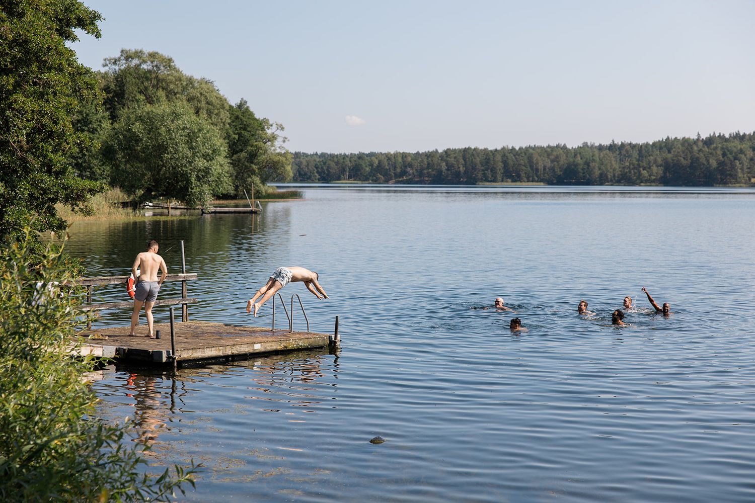 People swimming and enjoying a summer day at a peaceful lake with a wooden dock and forest shoreline.