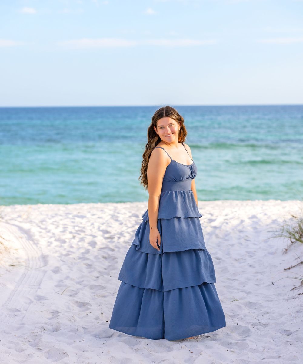 A person in a tiered blue maxi dress poses on a white sandy beach with turquoise ocean waters in the background.