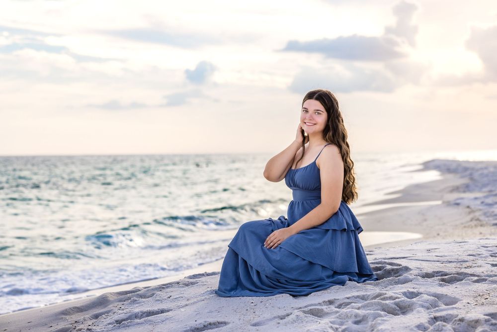 A person in a flowing blue dress poses gracefully along a white sandy beach at sunset.