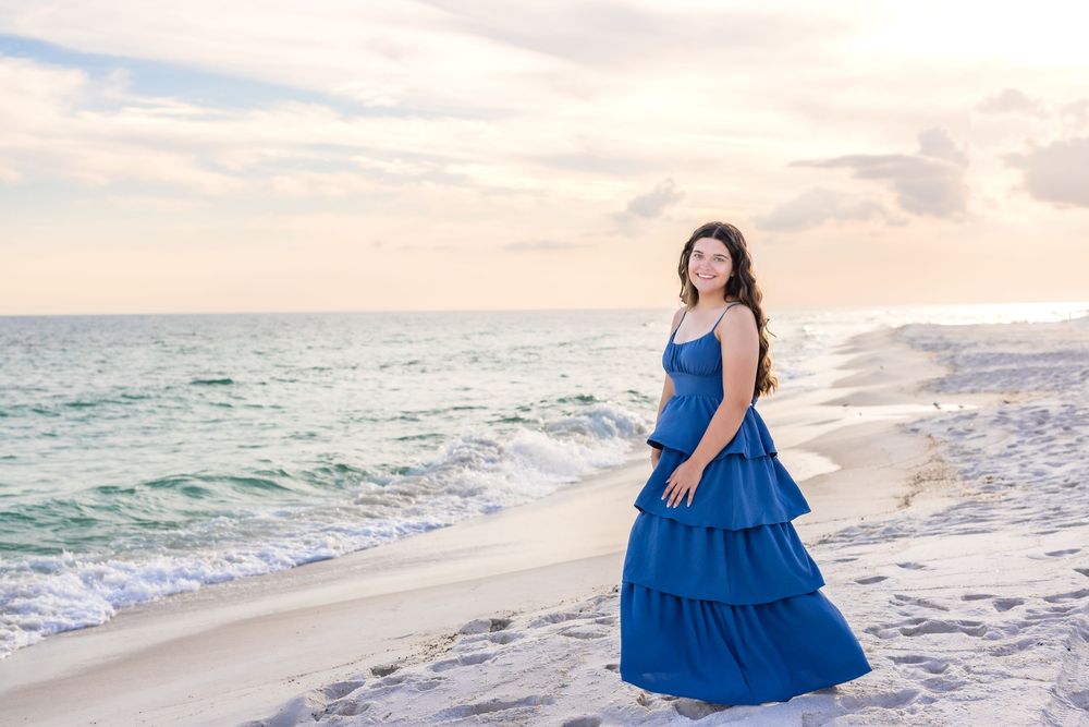 A figure in a flowing blue tiered dress stands on a white sandy beach at sunset with ocean waves in the background.