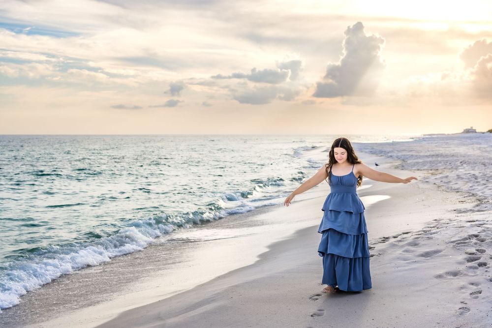 A scenic beach photoshoot at sunset showcasing someone in a flowing blue dress walking along the sandy shoreline.
