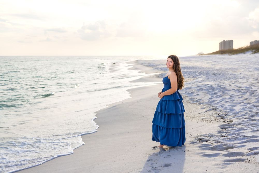 A person in a flowing blue dress walks along a pristine white sandy beach at sunset.