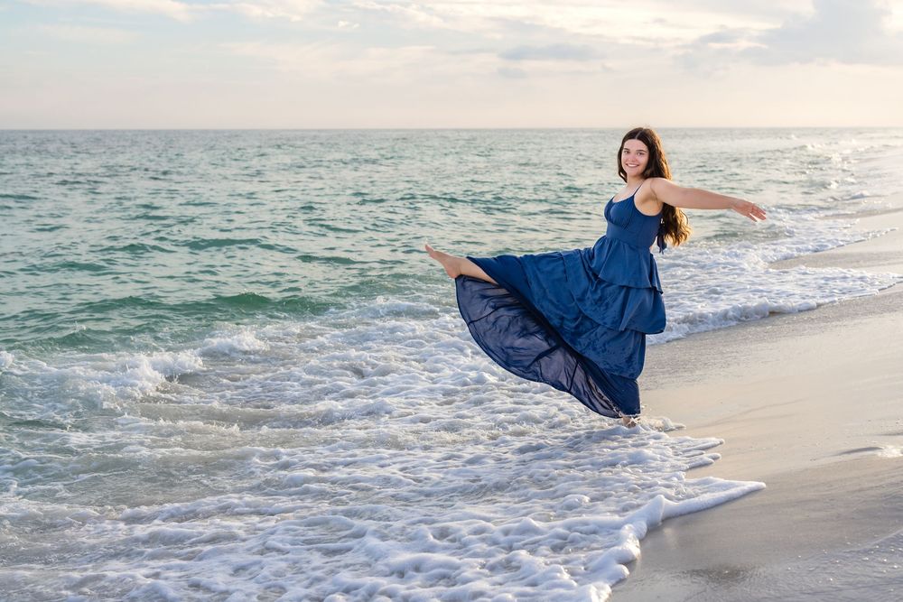 A person in a flowing blue dress dances gracefully along the ocean shoreline at sunset.