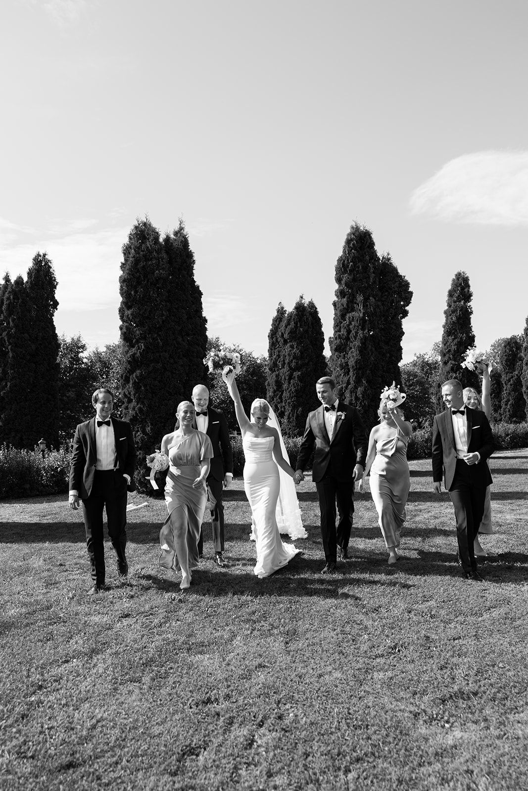 Black and white photo of a wedding party walking in a line across a grassy hill on a sunny day.