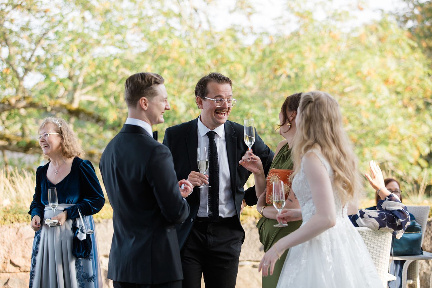 Wedding guests enjoying outdoor reception with laughter and conversation in natural autumn setting.