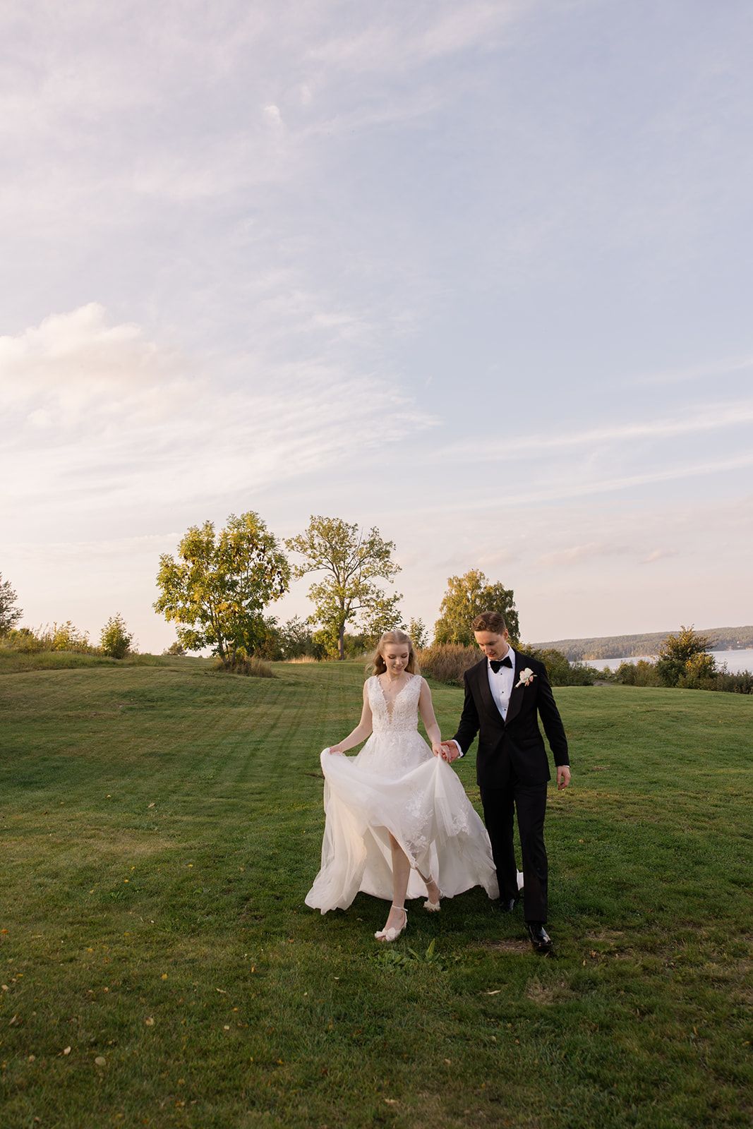 A couple in wedding attire walk hand in hand across a scenic hilltop meadow during golden hour.