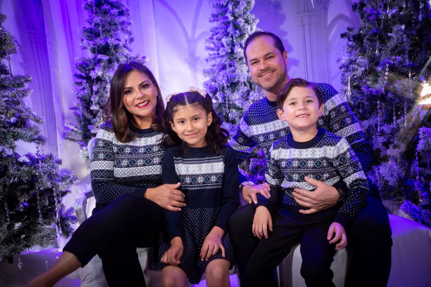 A group of people pose together at a festive holiday party with purple lighting and decorated Christmas trees.