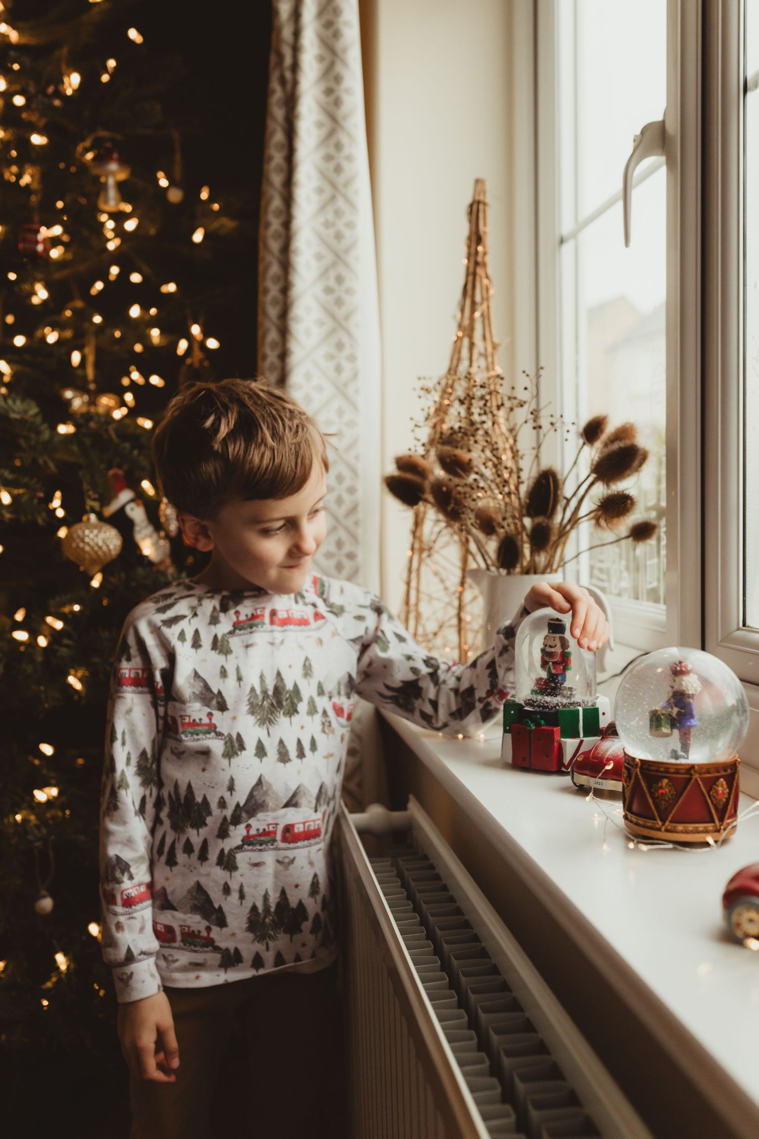 Child in holiday sweater gazes at Christmas decorations along windowsill with glowing tree lights in background.