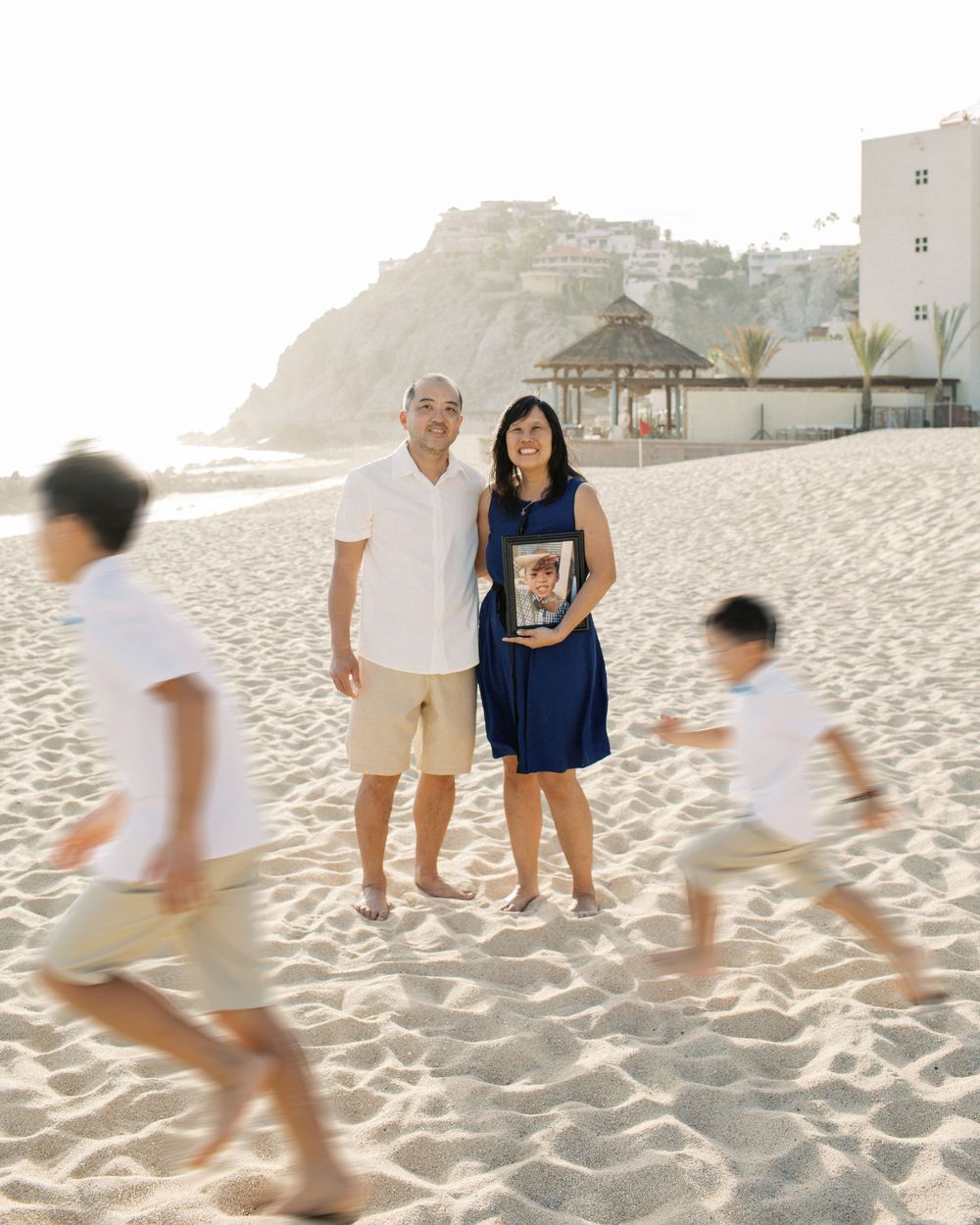 Family enjoying a sunny day at a sandy beach with gazebo and cliffs in the background as children run past.