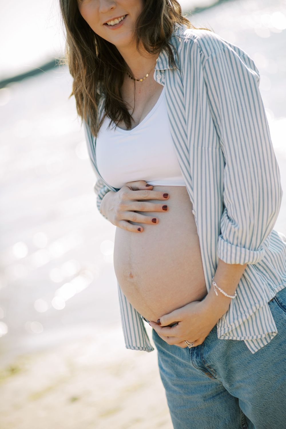 Expectant mother in striped shirt and jeans shows off baby bump in beach maternity photos.
