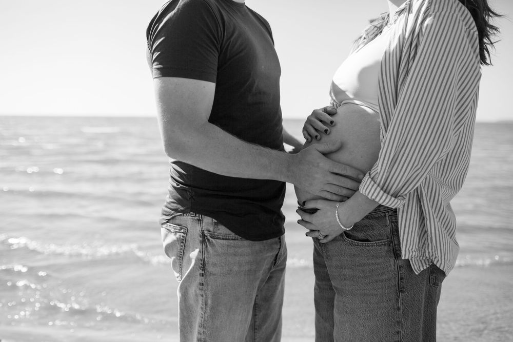 Intimate maternity photos of couple in casual wear against ocean backdrop.