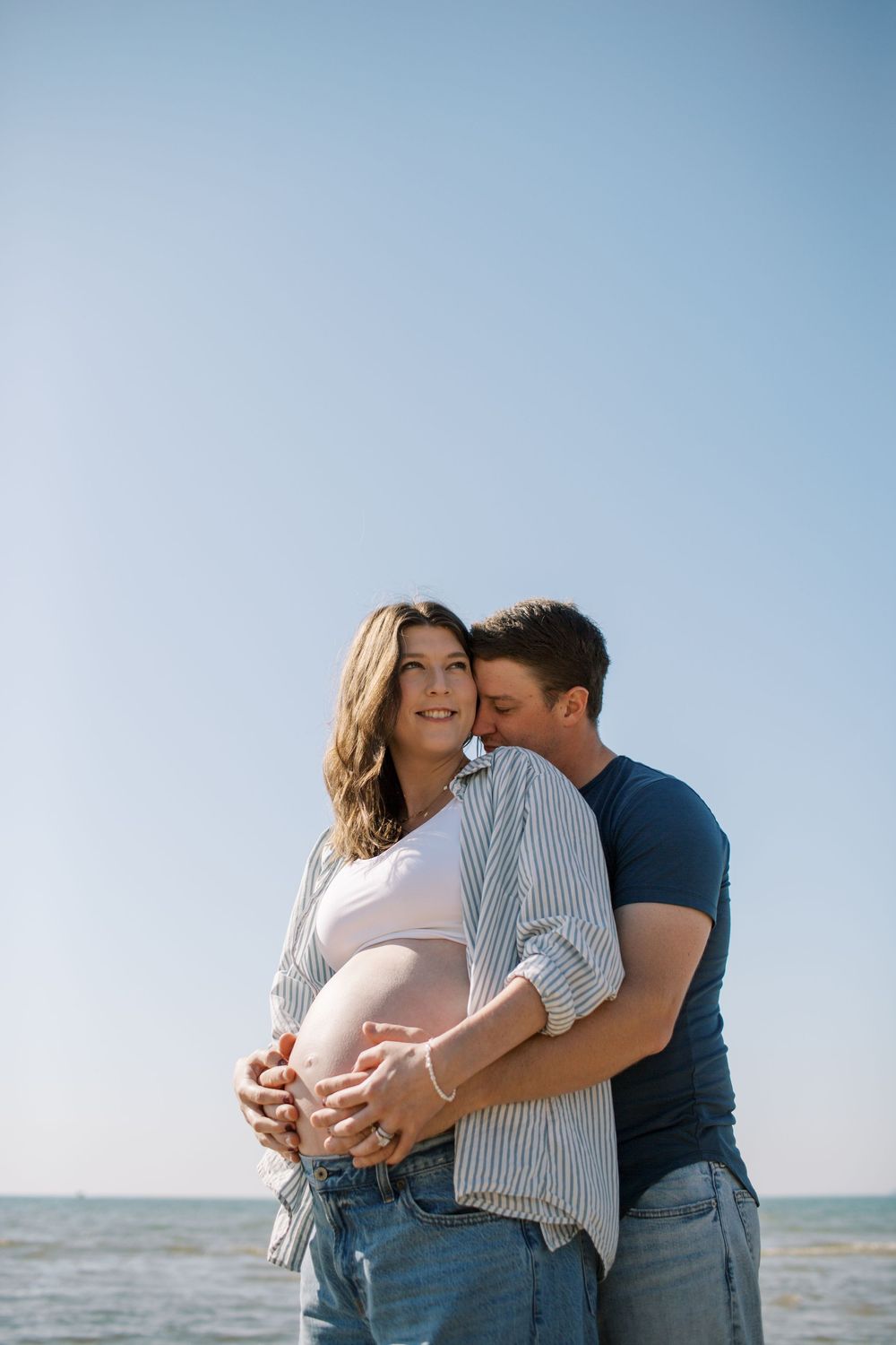 Maternity beach photoshoot featuring couple in casual clothes embracing against blue sky.