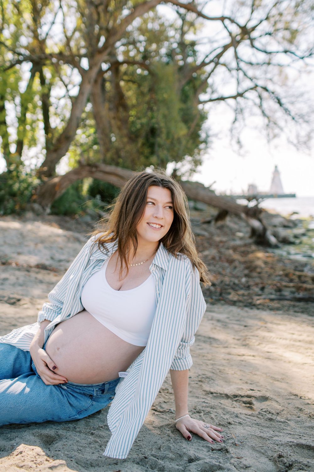 Expectant mother in striped shirt and jeans poses on beach with natural scenery in background.