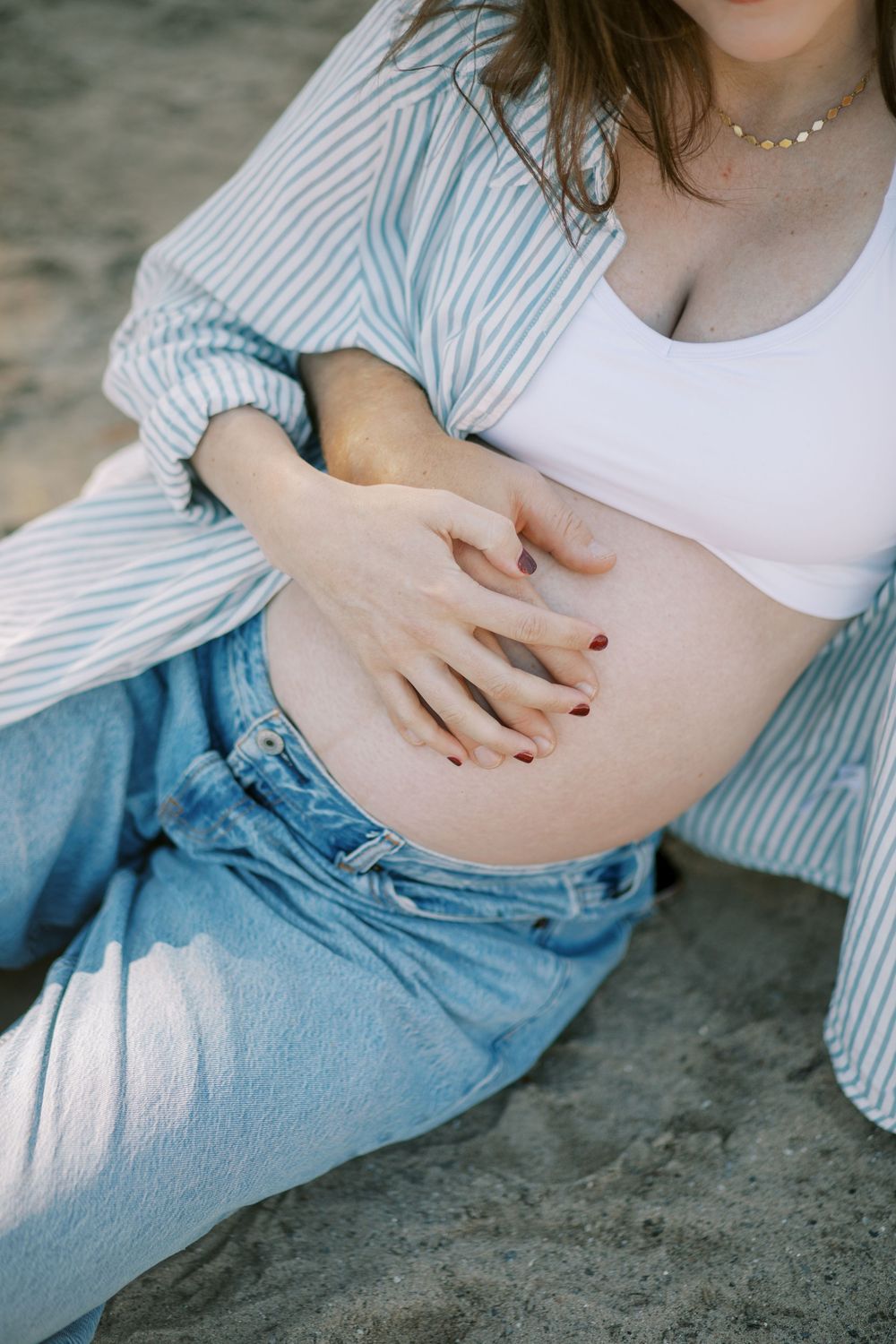 Close-up shot of pregnant belly with hands gently resting on it, wearing striped shirt and denim.