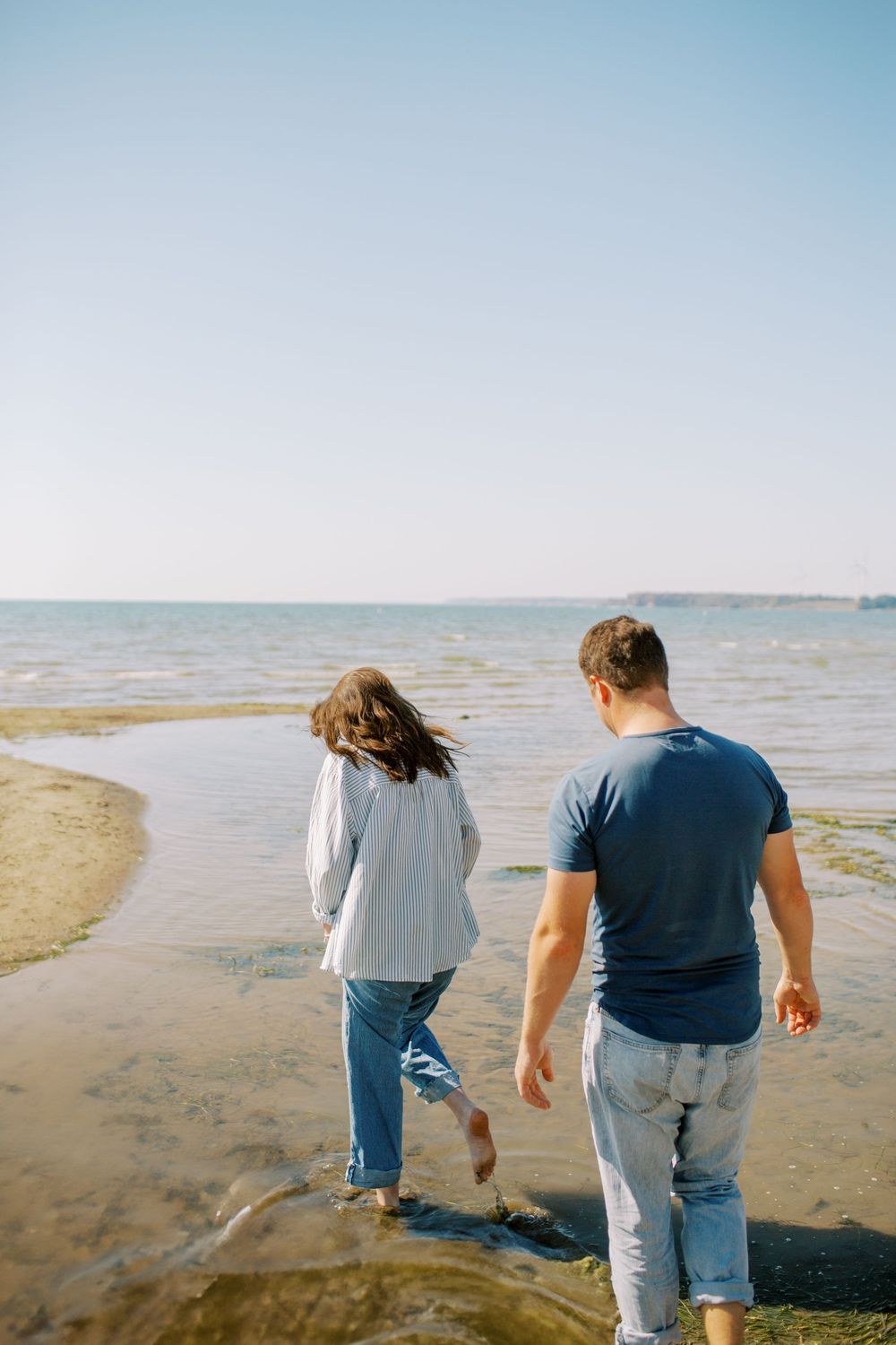 Couple walking along shoreline on a clear day with calm waters in the background.