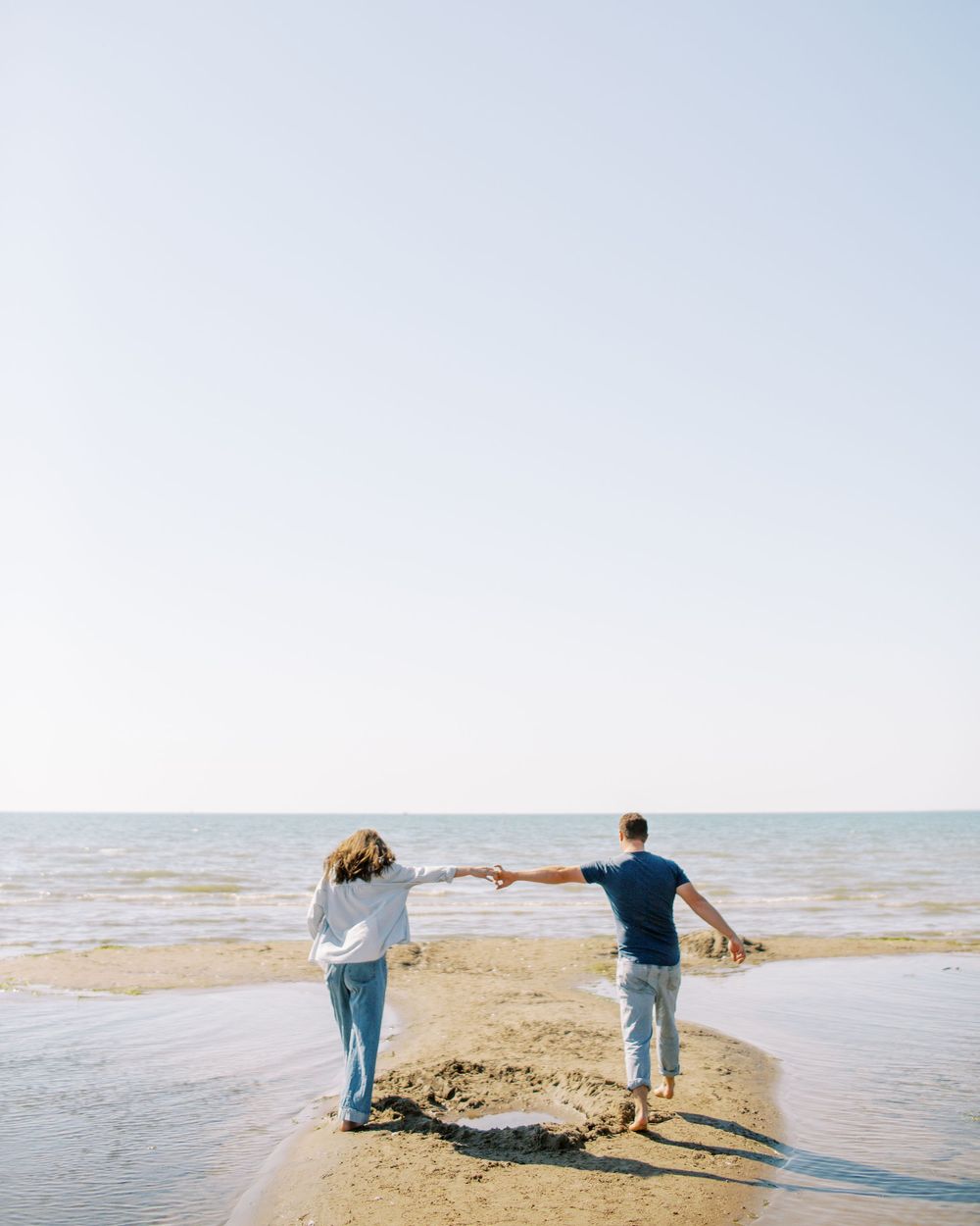 Couple holding hands while walking on a sandy beach peninsula extending into calm waters.