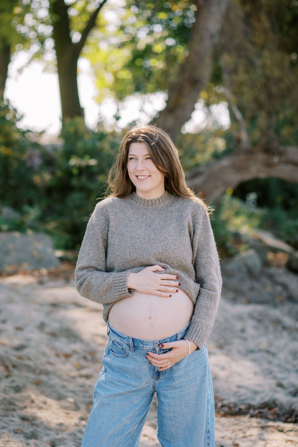 Pregnant woman in gray sweater and jeans showing her baby bump outdoors.