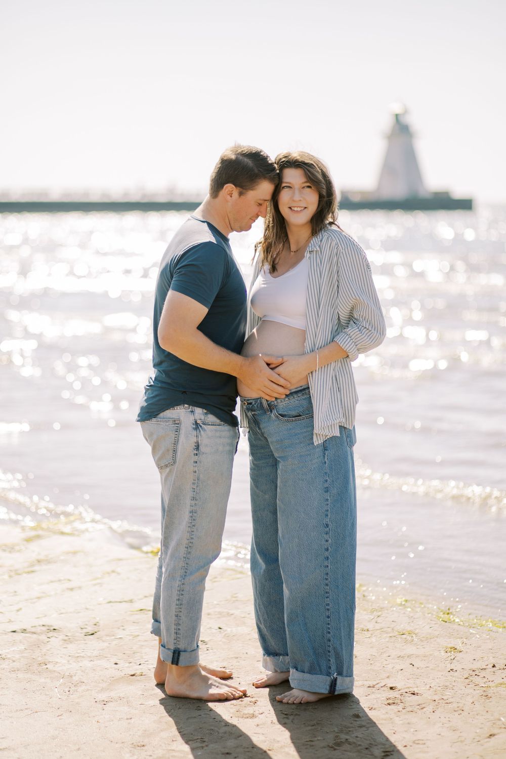 Couple in casual denim embracing on a sunlit beach during maternity photoshoot.
