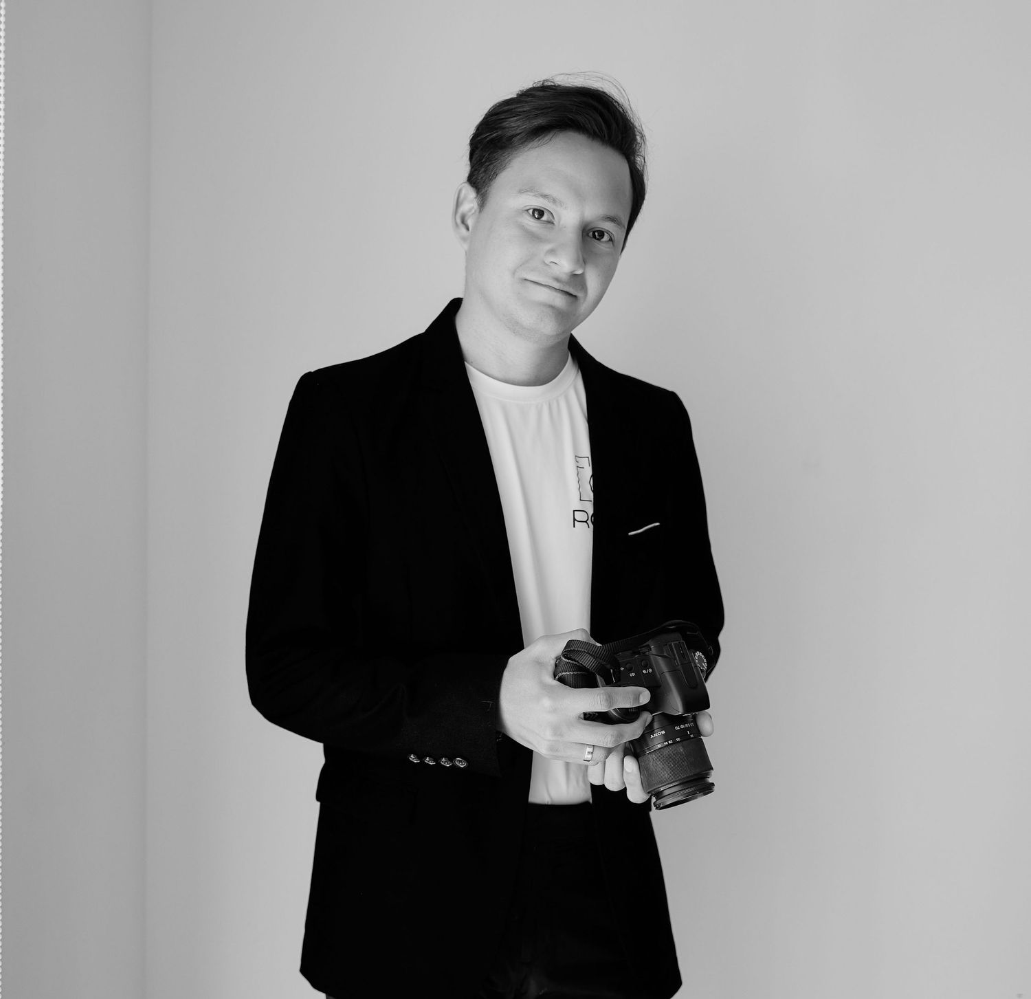 Professional in black blazer and white shirt holds camera while posing against plain wall in monochrome portrait.