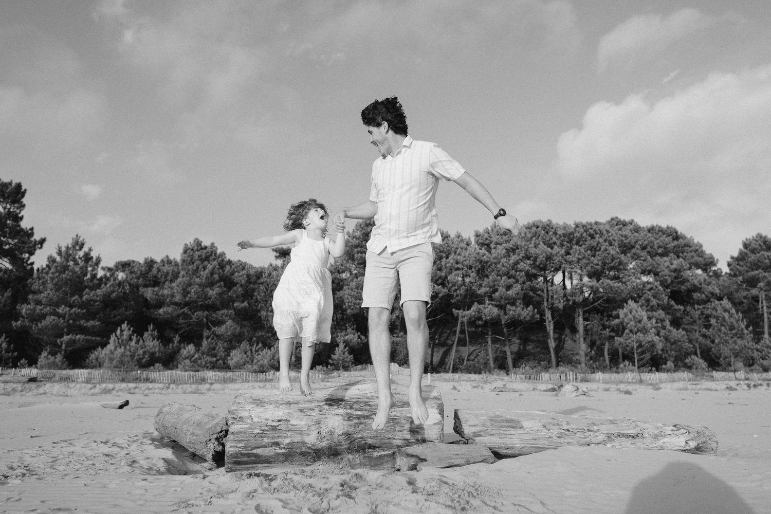 Un père et sa fille qui sautent par-dessus un tronc d'arbre se trouvant sur la plage.