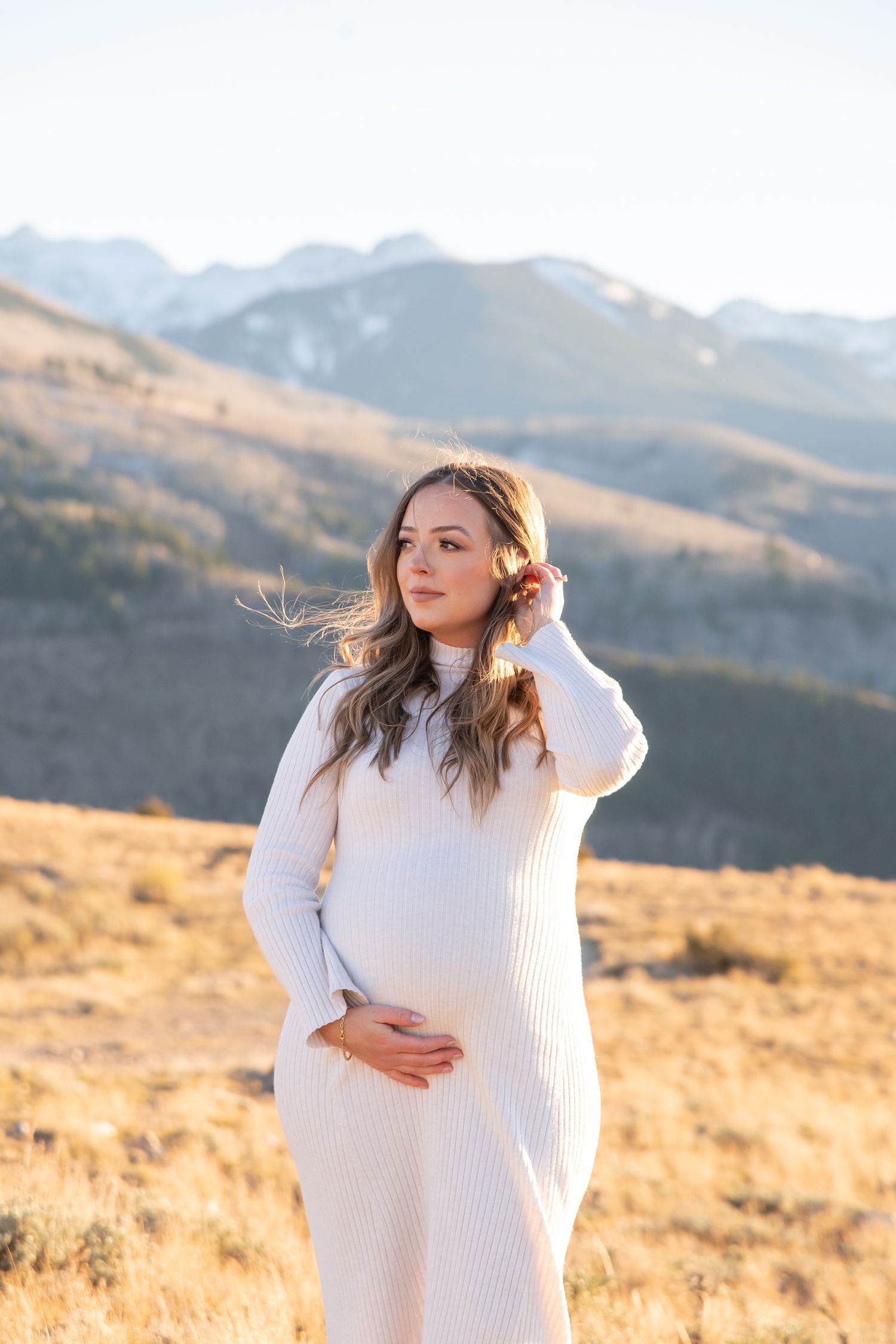 A beautiful woman models in a white dress and mountain peaks in the background during a maternity photo shoot in Vail Colorado.
