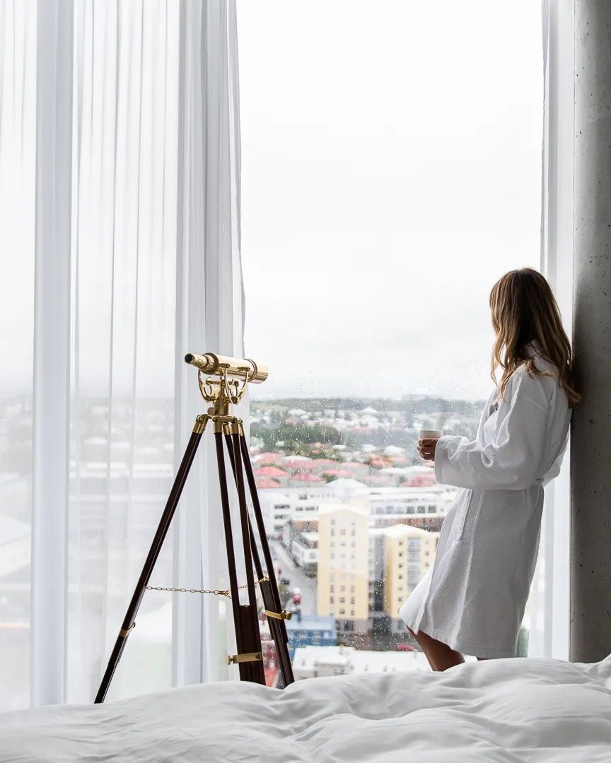 Woman standing by a large window beside a brass telescope overlooking the city.