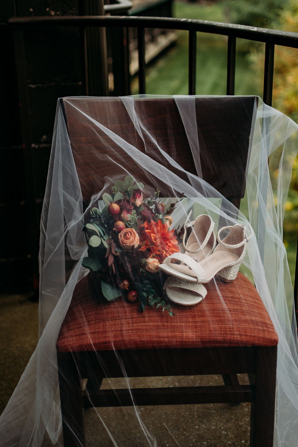 Wedding bouquet with pink roses and white heels arranged with delicate veil on a rustic wooden chair.