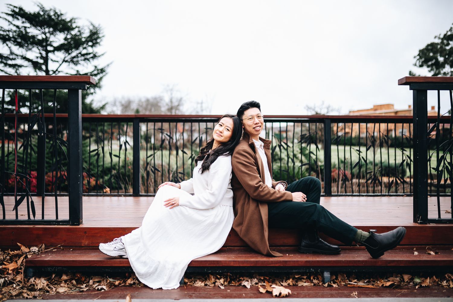 Two people in romantic autumn attire pose back-to-back at a park during a Melbourne engagement, prewedding and save the date photo session.
