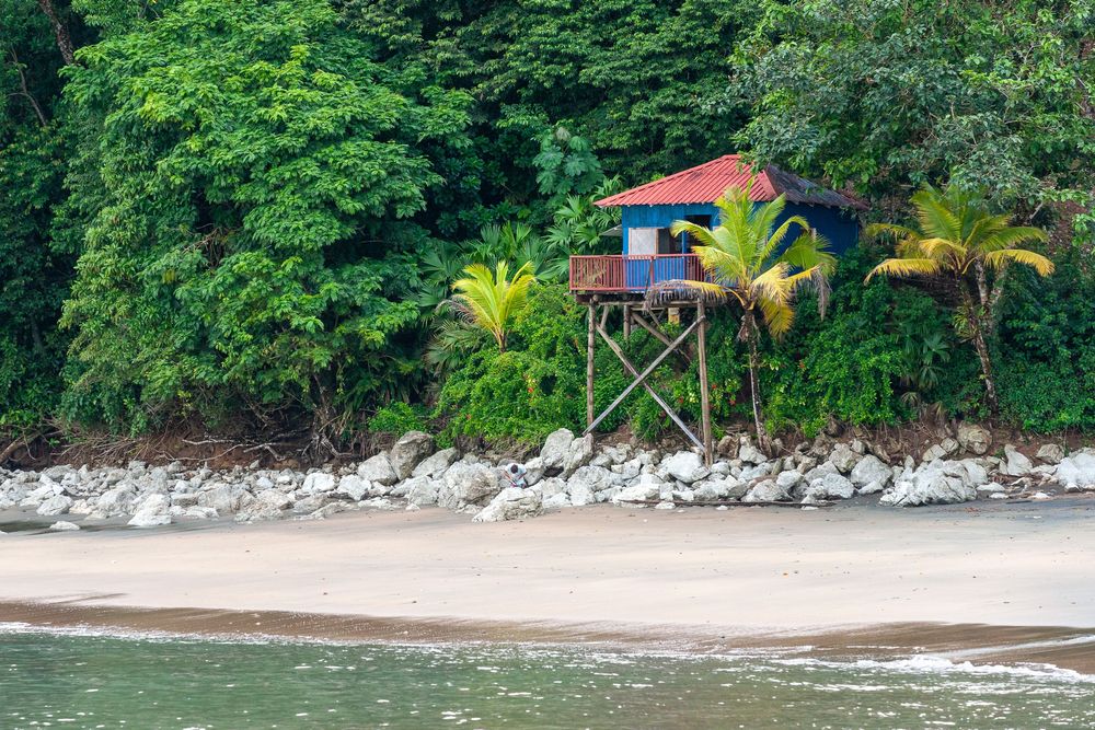 Colorful stilted beach hut with red roof perched above rocky shoreline among tropical palm trees and lush greenery.